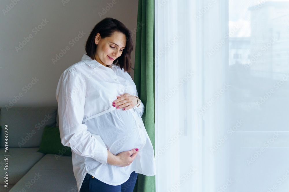 Smiling pregnant woman standing by window at home Smiling pregnant woman standing by window at home