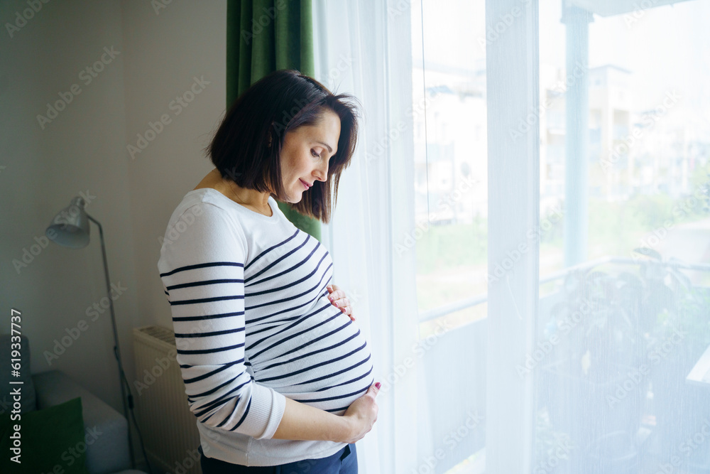 Smiling pregnant woman touching stomach and standing by window