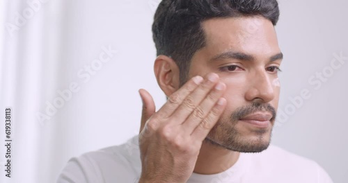 Handsome young man applying cream at his face while standing in front of mirror in bathroom at home.