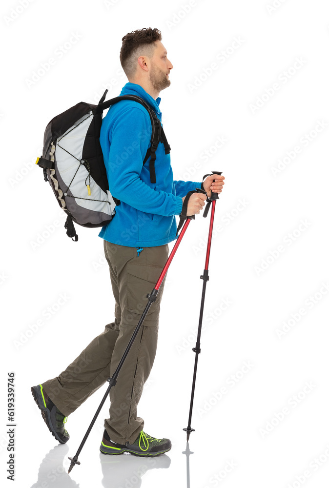 Full-length side view portrait of trekker with a backpack isolated on white background. Thirty years old man in blue jacket walking with trekking poles and posing in studio. Full-length side view portrait of trekker with a backpack isolated on white background. Thirty years old man in blue jacket walking with trekking poles and posing in studio.