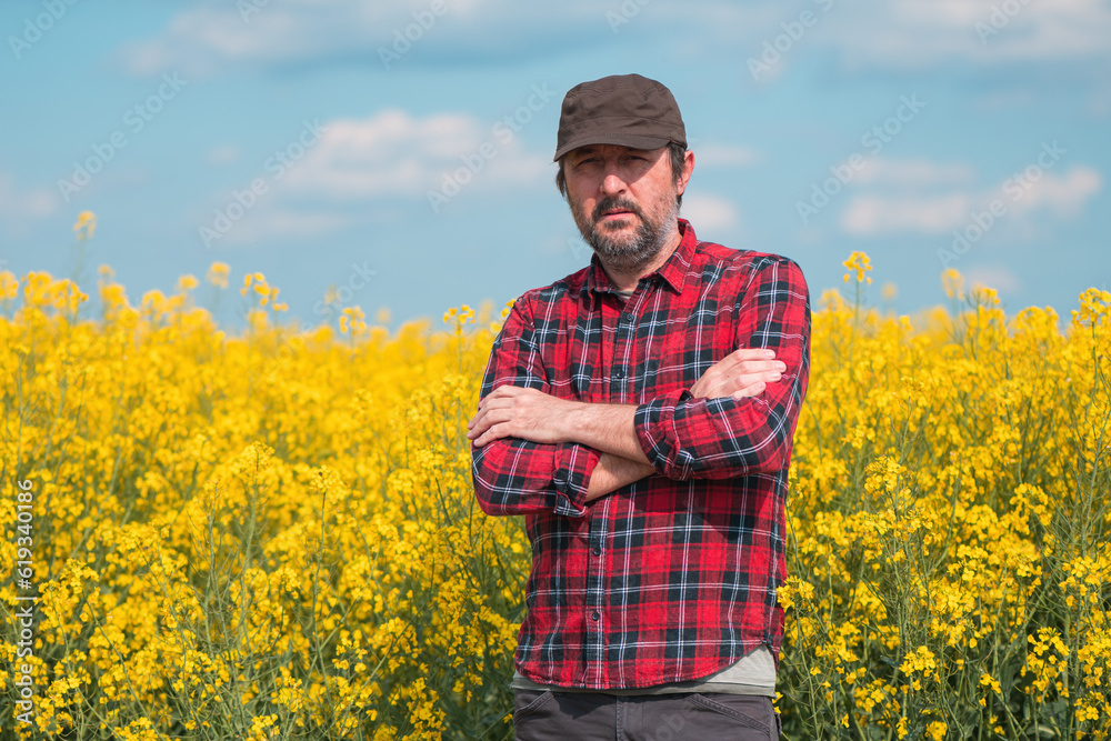 Portrait of confident farmer in blooming canola rapeseed field. Farm worker wearing red shirt and brown trucker's hat at plantation.