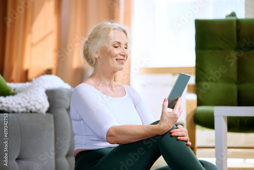 Beautiful, smiling, senior woman in sportswear looking on phone, watching online sport videos for training at home. Concept of sportive and healthy lifestyle, age, body care, fitness, wellness. © master1305