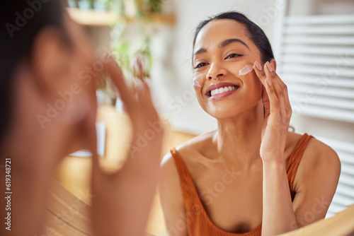 Smiling mixed race young woman applying moisturizer on her face in bathroom photo