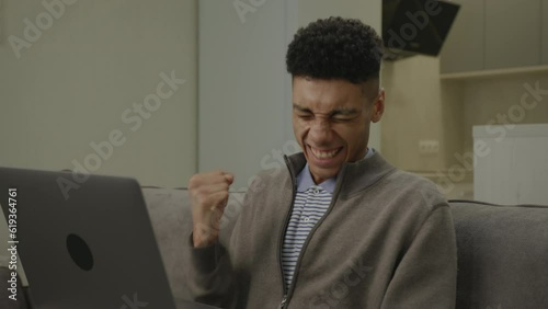 Man Receiving Grood News on Laptop. African American man is very happy using laptop sitting on sofa at home. photo