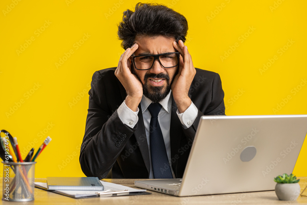 Tired ill indian businessman suffering from headache problem tension and migraine, stress at home office desk workplace. Bearded freelancer broker worker man guy works on laptop notebook. Overworking Tired ill indian businessman suffering from headache problem tension and migraine, stress at home office desk workplace. Bearded freelancer broker worker man guy works on laptop notebook. Overworking