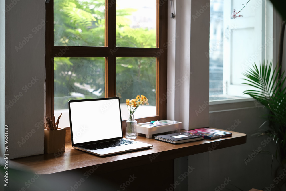 Comfortable home interior. Laptop with blank display, books and stationery on wooden desk near window