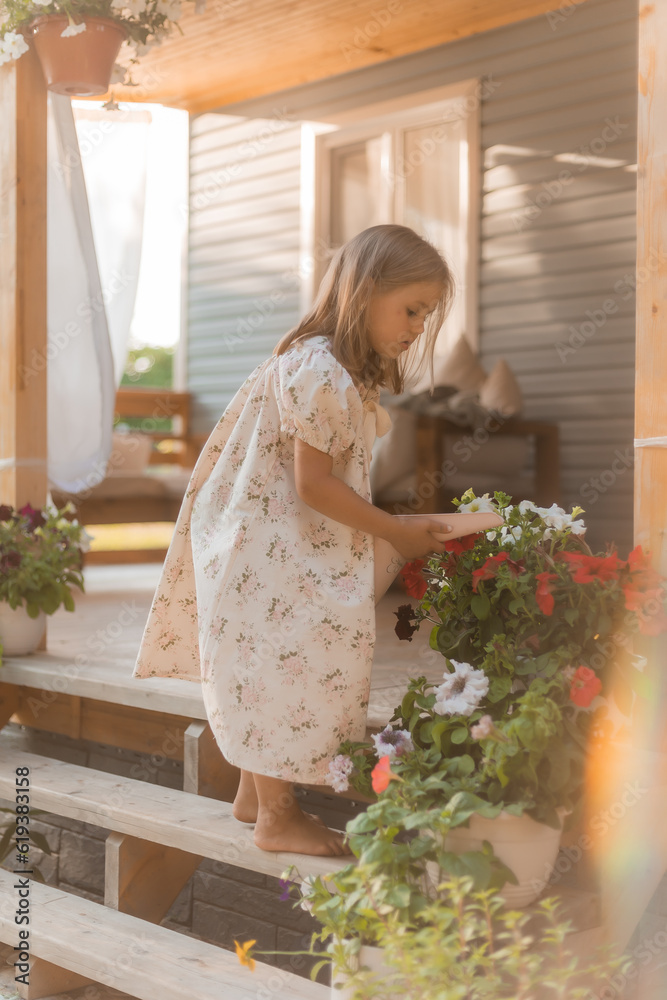  little girl with blonde hair is watering flowers on the veranda in summer at sunset