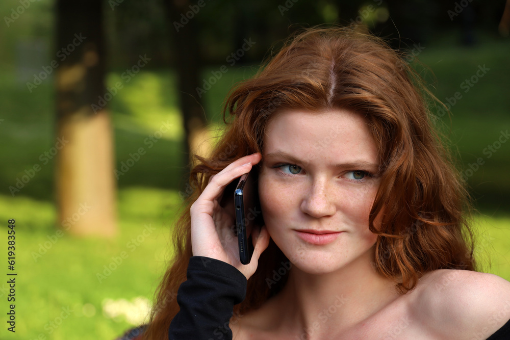 Smiling girl with long ginger hair and freckles talking on mobile phone sitting on a bench in summer park Smiling girl with long ginger hair and freckles talking on mobile phone sitting on a bench in summer park