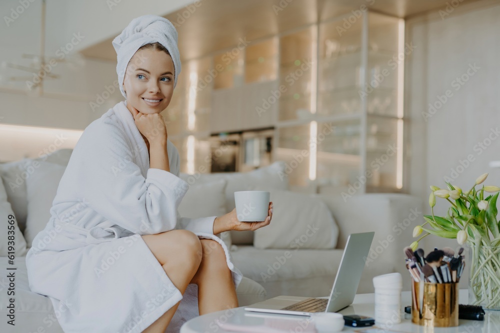 Cheerful woman in robe, fresh skin, drinks coffee, ponders, sits on sofa, works on laptop, uses cosmetics. Cheerful woman in robe, fresh skin, drinks coffee, ponders, sits on sofa, works on laptop, uses cosmetics.