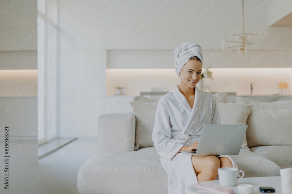 Pretty European woman works on laptop at home, in comfy clothes, on sofa, minimal makeup, smiling at camera. Pretty European woman works on laptop at home, in comfy clothes, on sofa, minimal makeup, smiling at camera.