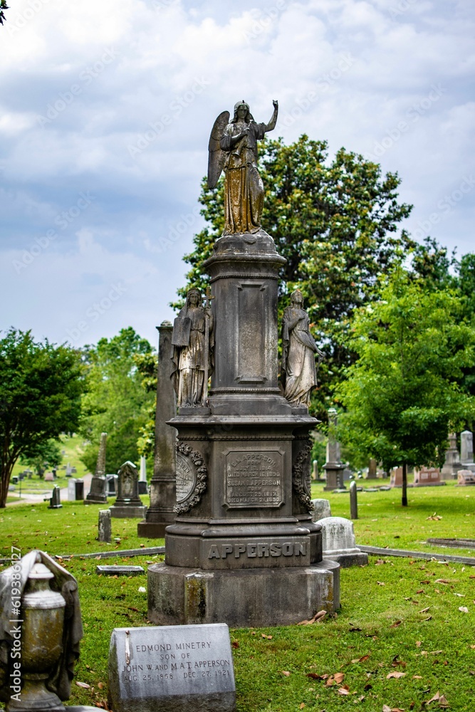 Statue atop a stone pedestal in a cemetery setting with a blurred background of graves and trees
