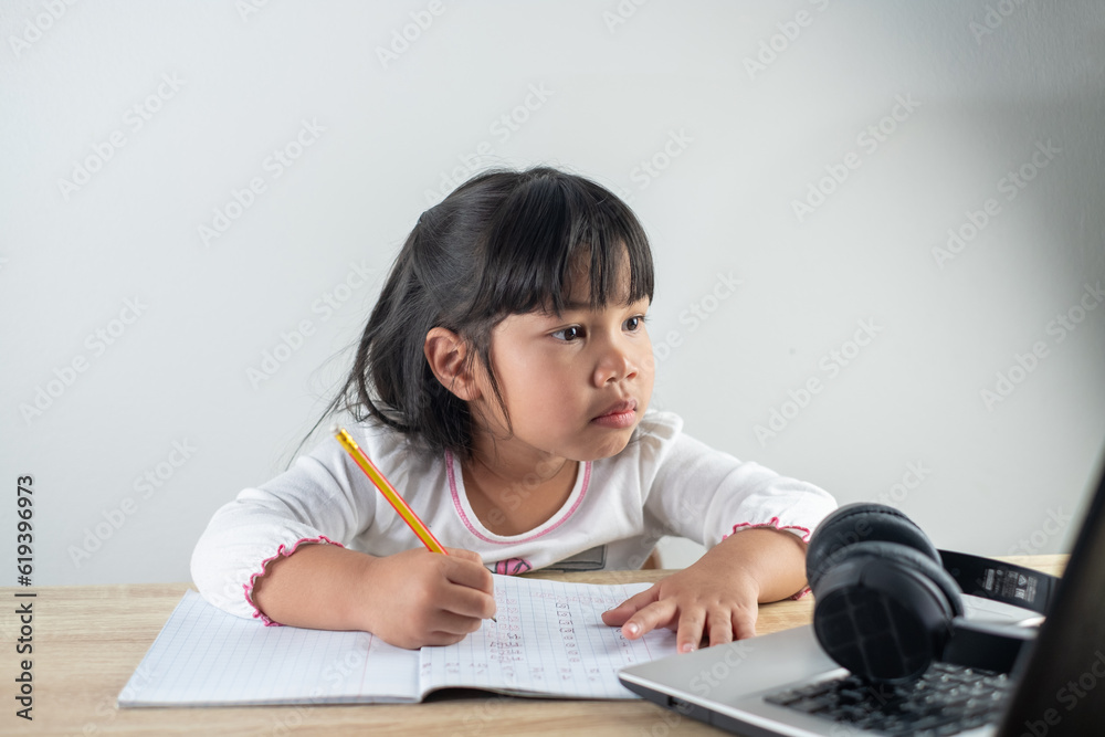 4-5 years old Asian girl wearing white shirt is studying online via computer using bluetooth headphones at home, distance education concept 4-5 years old Asian girl wearing white shirt is studying online via computer using bluetooth headphones at home, distance education concept