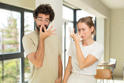 young adult couple feeling disgusted, holding nose to avoid smelling a foul and unpleasant stench photo