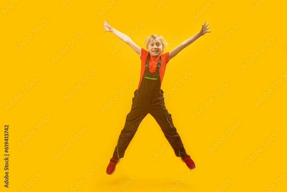 Portrait of schoolboy in overalls bouncing on yellow background. Blond boy jumps high with his hands up. Copy space