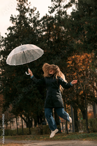 Cheerful student in black cloak with transparent umbrella jumps on alley of autumn park. Lush hair of girl covers face