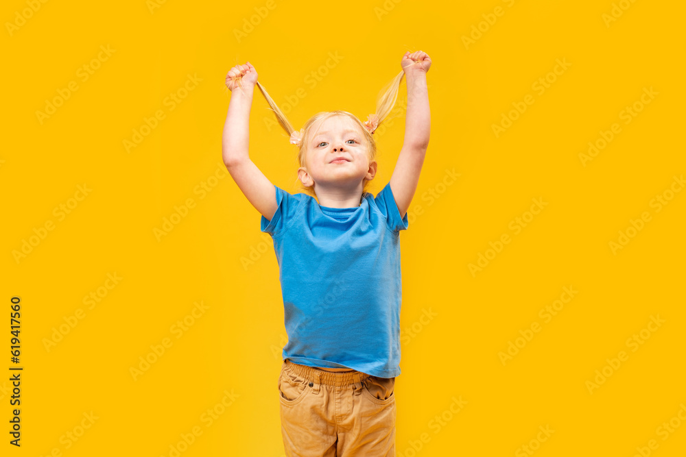 Portrait of smiling little girl having fun and holding two ponytails with her blonde hair. Yellow background Portrait of smiling little girl having fun and holding two ponytails with her blonde hair. Yellow background