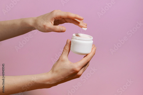 Female fragile hand takes cream from a white mockup jar which she holds on purple isolated background. The concept of beauty and aesthetics  natural cosmetics. Image for your design