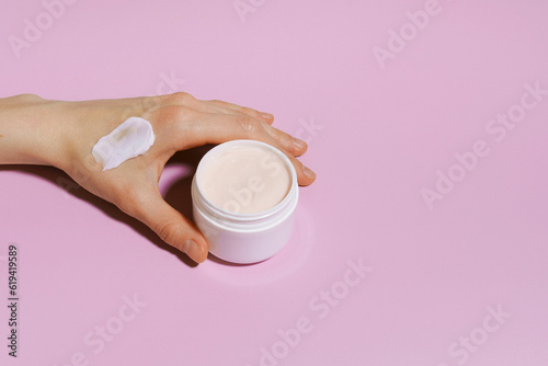 Female hand with a smear of pink face cream holds a jar on a purple isolated background. Image for your design. Beauty concept photo