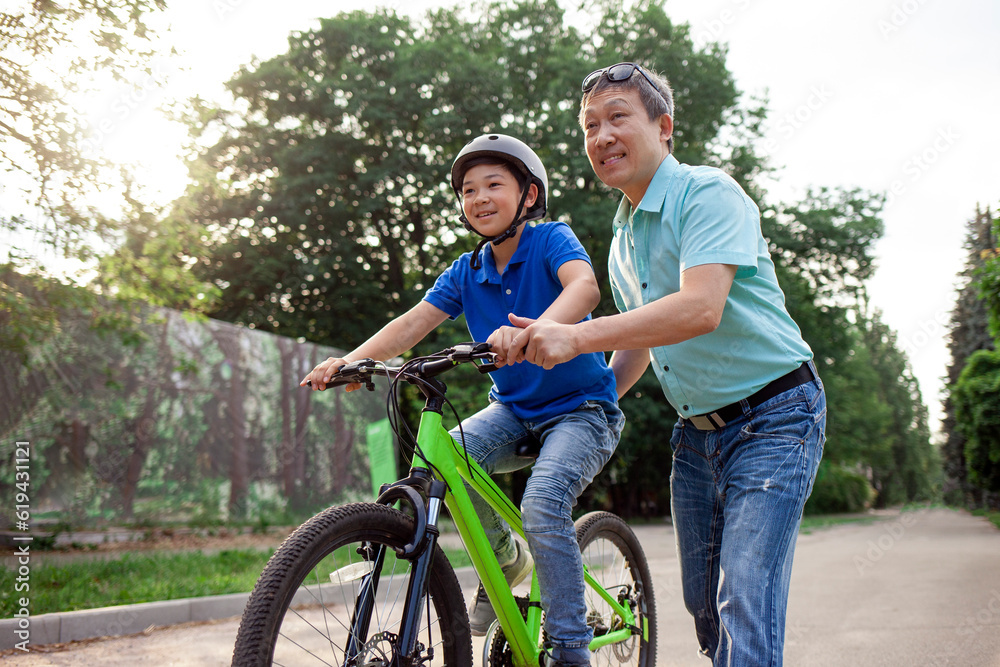 Obraz premium asian dad teaches son to ride bike, korean senior helps child, boy in helmet is actively relaxing with his father