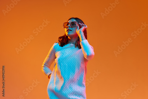 Portrait of young beautiful girl in casual clothes and trendy sunglasses posing against orange studio background in neon light. Concept of human emotions, youth, feelings, fashion, lifestyle, ad © master1305