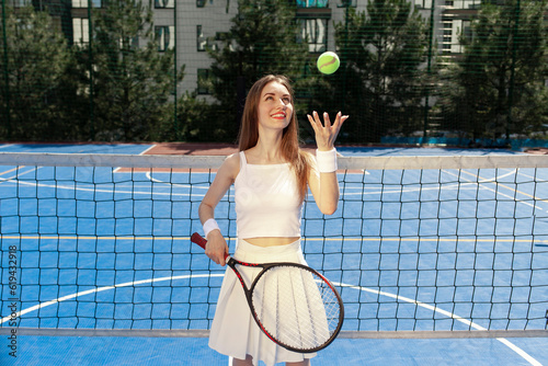 young girl tennis player in white uniform with racket throws up the ball on blue court outdoors