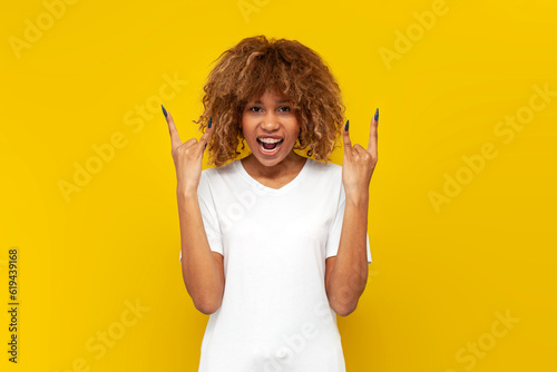 young curly american girl with braces screaming and showing rock gesture on yellow isolated background