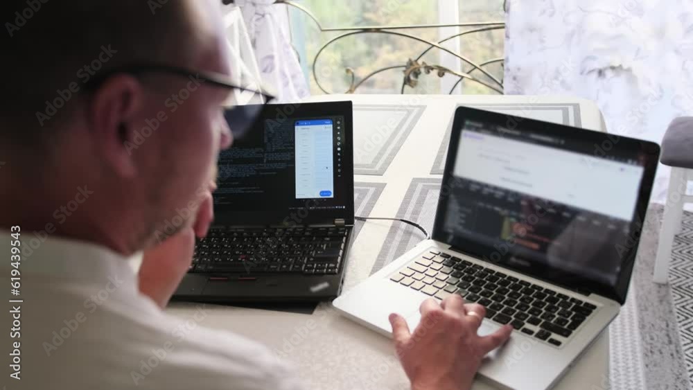 Focused IT specialist in glasses writes program code on two laptops while working at home near the window overlooking street, rear view. Programmer work in home office. Remote work arrangements. Focused IT specialist in glasses writes program code on two laptops while working at home near the window overlooking street, rear view. Programmer work in home office. Remote work arrangements.