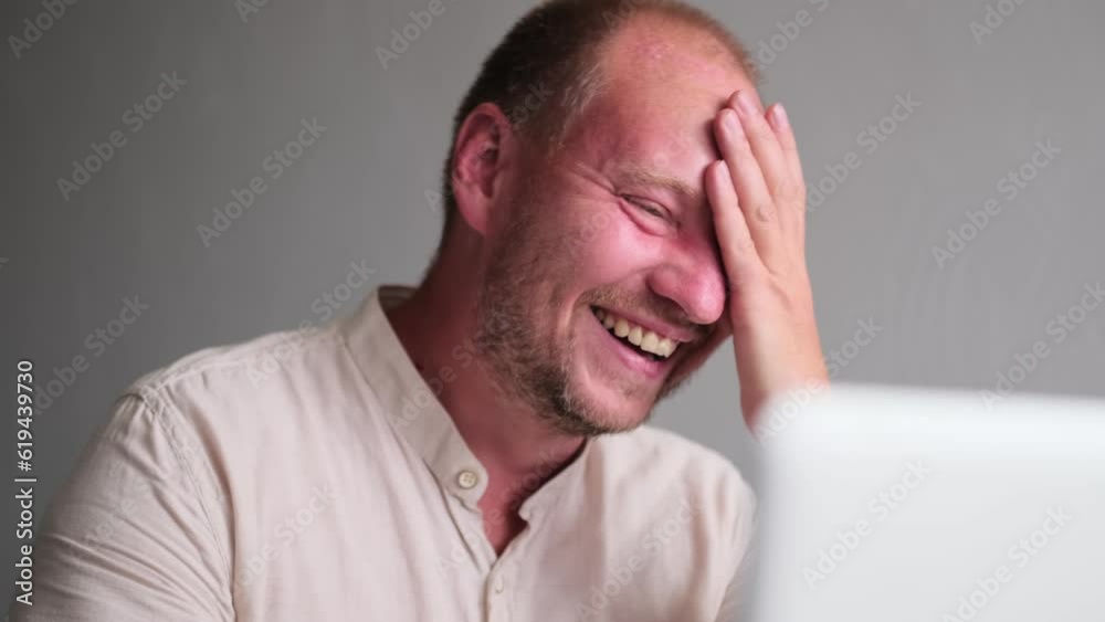 Adult man in a good mood laughs looking at the monitor screen against a light wall, close up. Caucasian man smiles in a break between work, wearily rubbing his unshaven face. Adult man in a good mood laughs looking at the monitor screen against a light wall, close up. Caucasian man smiles in a break between work, wearily rubbing his unshaven face.
