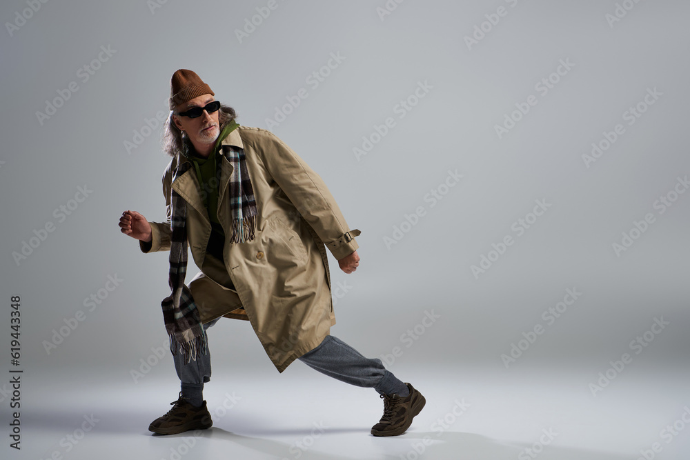 full length of fashionable senior man looking away on grey background, stylish pose, hipster, dark sunglasses, beanie hat, beige trench coat, stylish aging of expressive personality