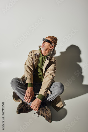 full length of senior hipster man in beanie hat, dark sunglasses and beige trench coat sitting with crossed legs and looking away on grey background with shadow, fashionable aging concept