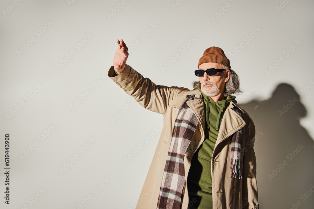 fashionable senior man in dark sunglasses, beanie hat, beige trench coat and plaid scarf standing with outstretched and looking away on grey background, hipster style, expressive personality fashionable senior man in dark sunglasses, beanie hat, beige trench coat and plaid scarf standing with outstretched and looking away on grey background, hipster style, expressive personality