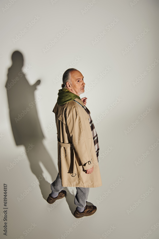 full length of thoughtful senior man in green hoodie, beige trench coat and plaid blazer looking away on grey background with shadow, fashionable lifestyle, high angle view full length of thoughtful senior man in green hoodie, beige trench coat and plaid blazer looking away on grey background with shadow, fashionable lifestyle, high angle view