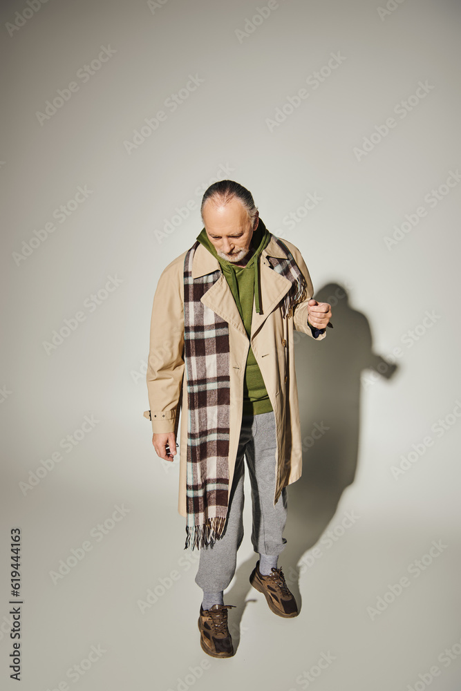 full length of aged bearded man looking down while standing in trendy casual attire on grey background with shadow, beige trench coat, plaid scarf, green hoodie, fashion and age concept full length of aged bearded man looking down while standing in trendy casual attire on grey background with shadow, beige trench coat, plaid scarf, green hoodie, fashion and age concept