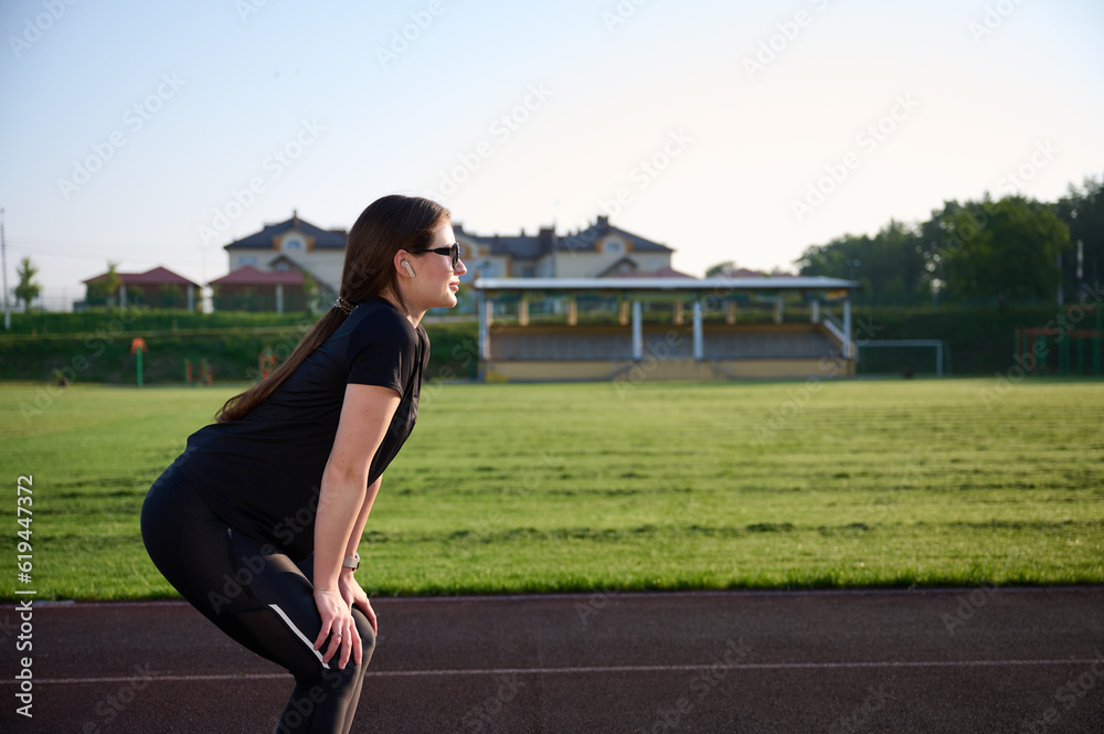Portrait of young attractive fit girl in black sports tight clothes standing on green field before workout at sunrise. Caucasian female jogger runner on jogging track. Sport and health. Portrait of young attractive fit girl in black sports tight clothes standing on green field before workout at sunrise. Caucasian female jogger runner on jogging track. Sport and health.