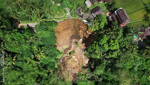 Aerial camera provides an overview of landslide aftermath, then descends to capture detailed view and cave at bottom of fresh cliff. At top, buildings perilously stand on edge of precipice photo