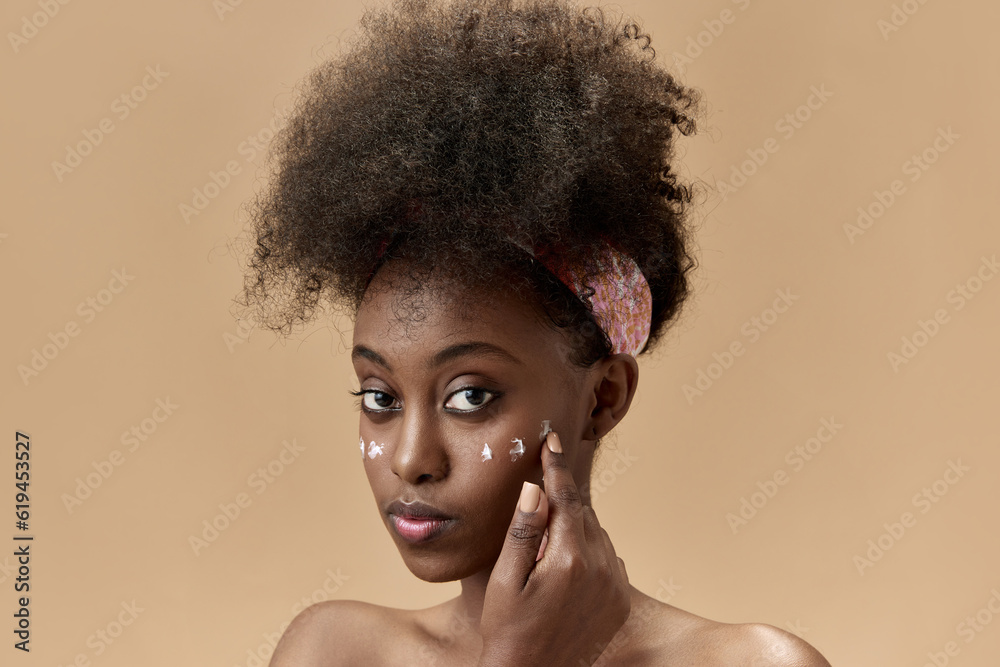 Close-up portrait of young, beautiful, african woman applying face moisturizing cream against studio background Close-up portrait of young, beautiful, african woman applying face moisturizing cream against studio background
