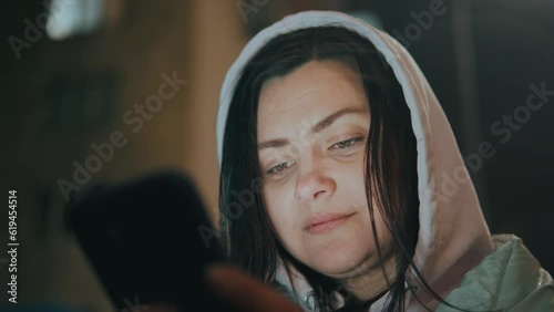 Girl's face illuminated by a smartphone screen in a hood in the rain at night
