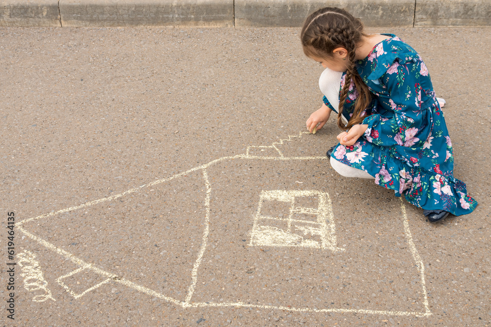Naklejka premium Junior school girl draws windows and curtains of house with white chalk on asphalt. Girl in flowery dress enjoys drawing house on asphalt, side view
