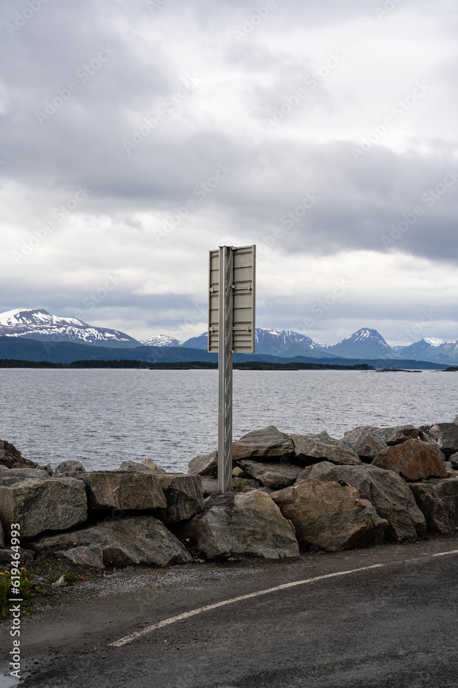 A Norwegian fjord on a harsh overcast day with dark fjords in the distance with white snow capped with a dramatic sky and rocks and a road sign in the foreground