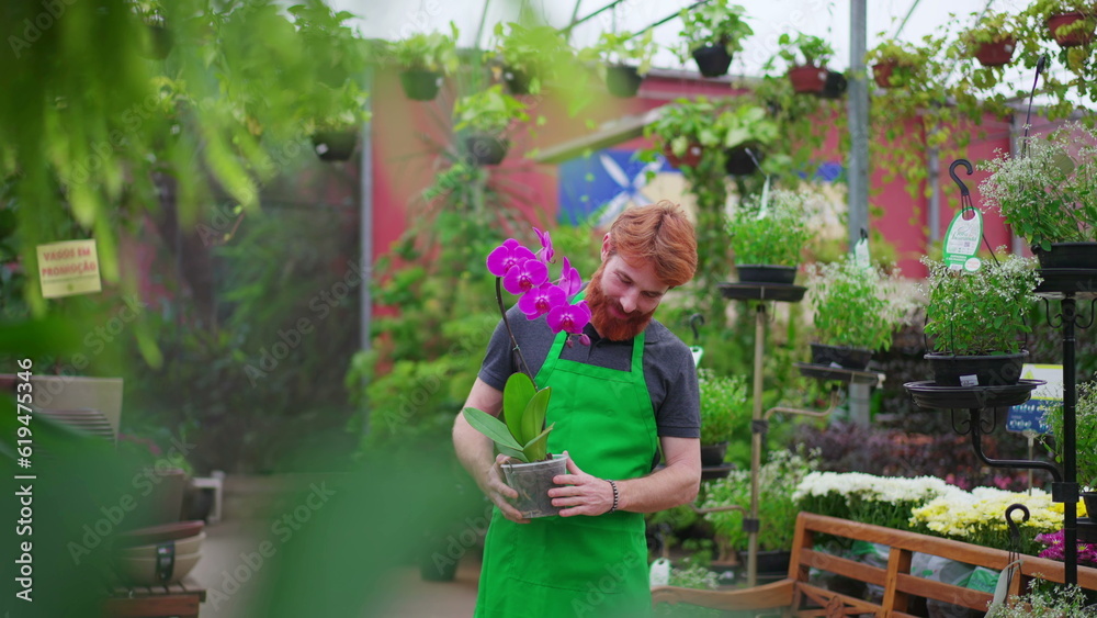 Smiling Young Man with Flower at Horticulture Shop. Redhead Employee in Green Apron in Local Store