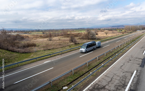 A modern gray minibus is driving, with a hitched trailer for transporting things on the highway.