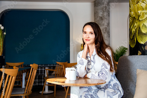 Beautiful young woman with long brown hair sitting in cafe and dreaming about meeting. Brunette with happy face is smiling and waiting. Expectation concept  © Yelena