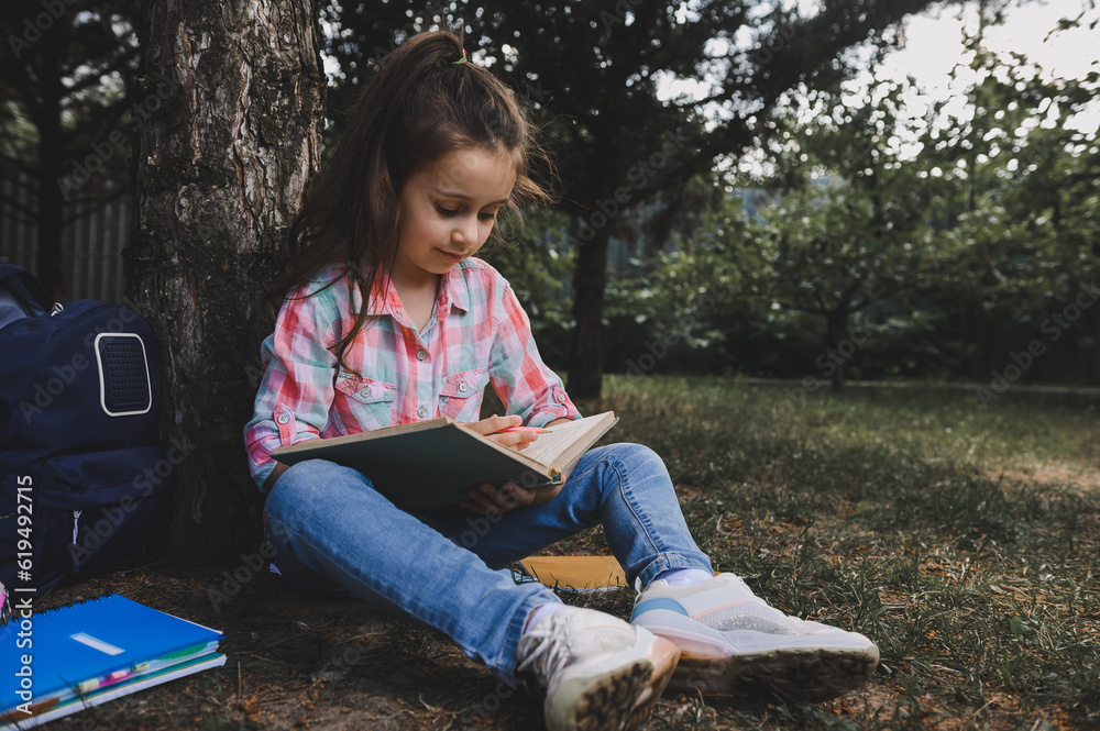Beautiful diligent little child, primary school student girl, sitting under a tree in the park, reading a book. Beautiful diligent little child, primary school student girl, sitting under a tree in the park, reading a book.