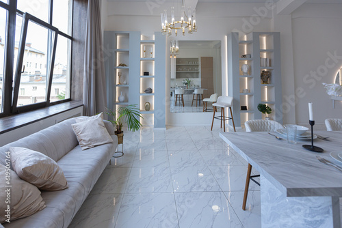 minimalistic light elegant luxury design of a modern spacious studio apartment with kitchen area, bedroom and huge mirror behind the couch. tiles on the floor and high ceiling.