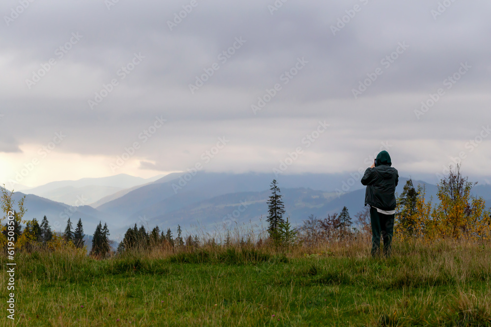 Guy in jacket and pants takes pictures of beautiful autumn cloudy mountains with his phone. Beautiful autumn landscape, sky in clouds, grass and trees. Tourism and travel