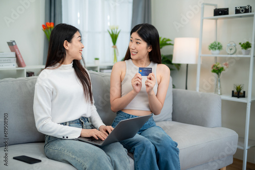 Two young Asian women express their surprise while using their credit card to make purchases. Successful online shopping with a laptop on the sofa in the living room.