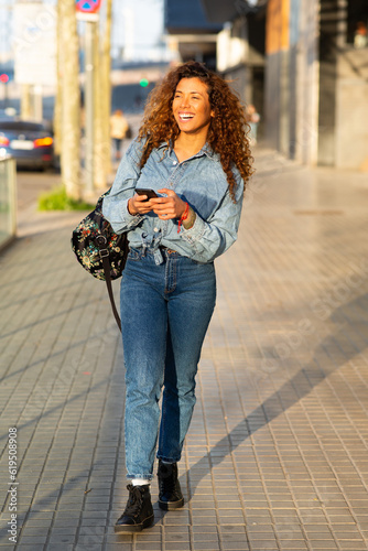 Smiling young latin woman walking with bag and mobile phone outside in the city