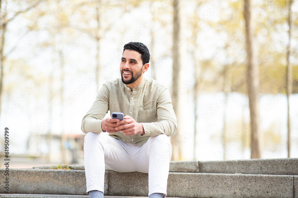 Fototapeta premium Young middle eastern guy sitting outside with mobile phone