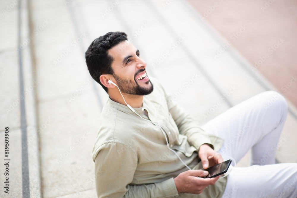 Smiling arabic man sitting outside enjoying listening to music from his cell phone