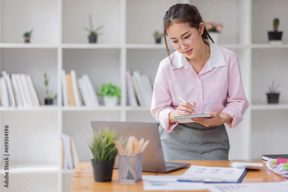 Asian businesswoman working on a laptop computer at her desk in a bright modern office, doing calculating expense financial report finance making notes paper graph data document. Asian businesswoman working on a laptop computer at her desk in a bright modern office, doing calculating expense financial report finance making notes paper graph data document.